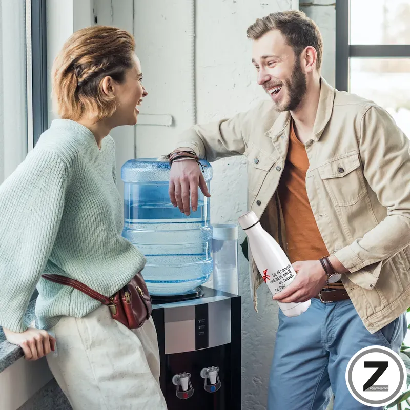 Two coworkers chat by a water cooler, smiling and relaxed in a bright office break area. The man holds a bottle while the woman leans on the counter.