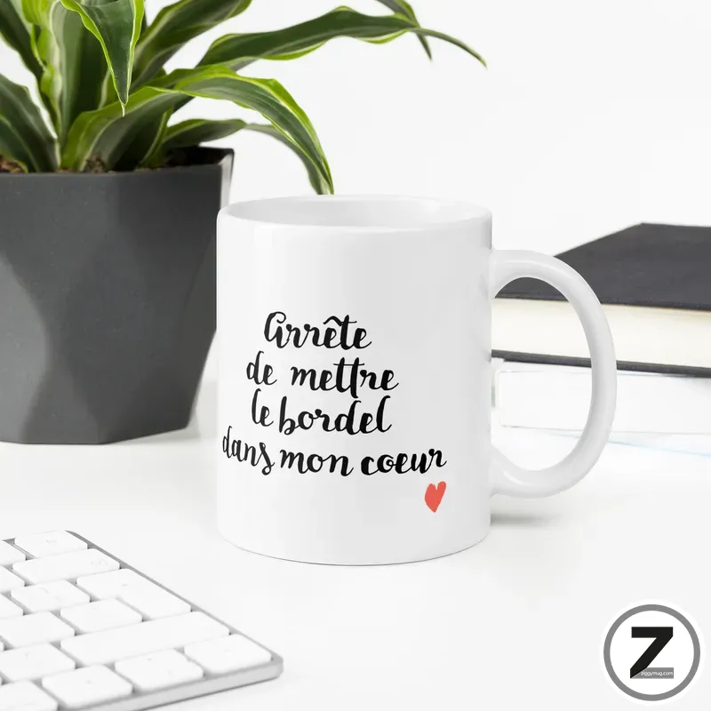 White mug on a desk with the French phrase “Arrête de mettre le bordel dans mon cœur” and a small red heart, with a plant in the background.