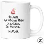 White mug with a French birthday joke: '19 ans, ça mérite bien un cadeau de merde. Un mug.' and a small party hat.