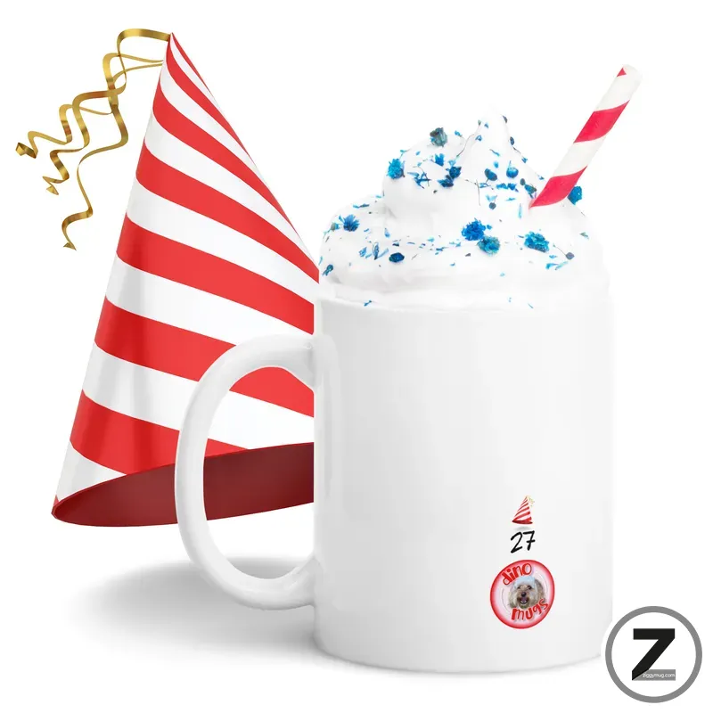 White ceramic mug filled with whipped cream and blue sprinkles, with a red-and-white striped straw; a matching red-and-white striped party hat leans beside the mug, festive ribbons curling nearby; a small sticker/logo is on the mug's side.