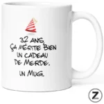 Mug blanc affichant le texte: '32 ans ça mérite bien un cadeau de merde. un mug.' avec un petit chapeau de fête sur l’image.