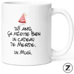 White ceramic mug featuring a red party hat and a humorous French message about turning 38: 'ça mérite bien un cadeau de merde. Un mug.'