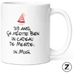 White ceramic mug featuring a humorous French phrase in black handwritten font and a small red-and-white party hat graphic. The text reads: '39 ans, ça mérite bien un cadeau de merde. Un mug.'