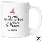 White ceramic mug featuring a French humorous message about turning 43 with a small party hat illustration above the text.