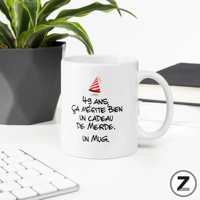 White ceramic mug on a desk with a plant and keyboard, featuring a French joke and a small red party hat graphic.