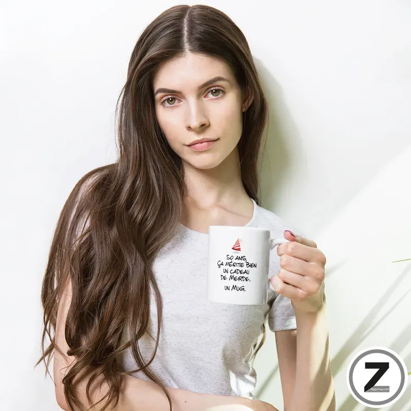 Young woman with long brown hair wearing a light gray t‑shirt, holding a white mug with French text, standing in bright light.