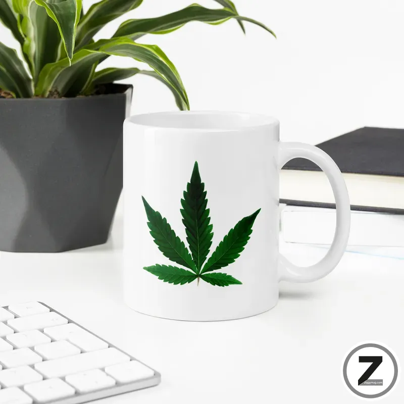 White ceramic mug with a large green cannabis leaf design on a bright desk next to a plant pot and keyboard.