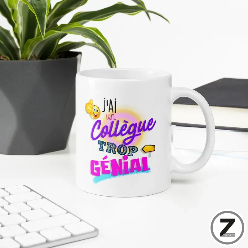 White mug on a desk with colorful text saying "J'ai un collègue trop génial" and a small emoji, next to a plant and keyboard in a bright office setting.