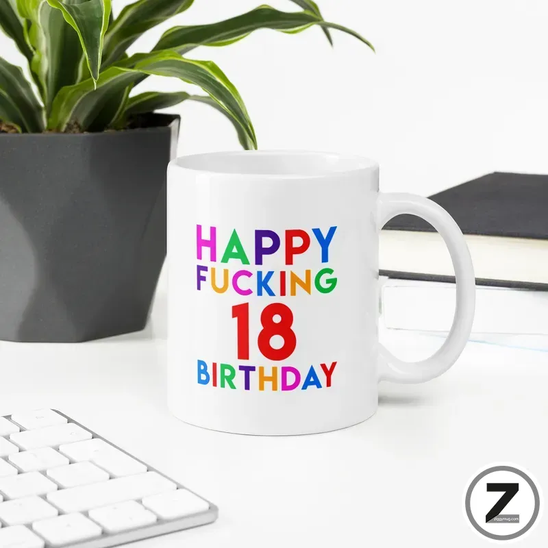 White ceramic mug with rainbow text reading "HAPPY FUCKING 18 BIRTHDAY" on a desk beside a plant and keyboard.