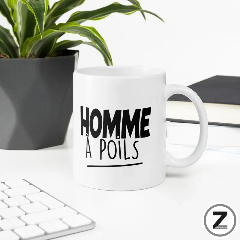 White ceramic mug on a desk with the bold black text 'HOMME A POILS' printed on it, beside a plant and books.