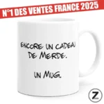 White mug with handwritten French text 'Encore un cadeau de merde. Un mug.' A red promo banner across the top reads 'N°1 DES VENTES FRANCE 2025'.
