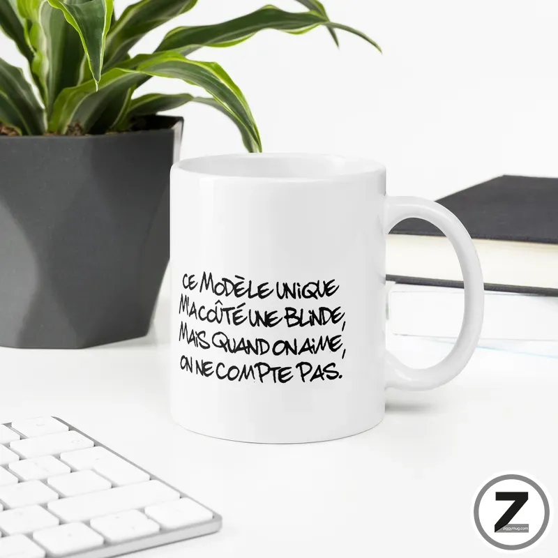 White ceramic mug on a desk with a bold handwritten French slogan, next to a potted plant and keyboard in a bright workspace.