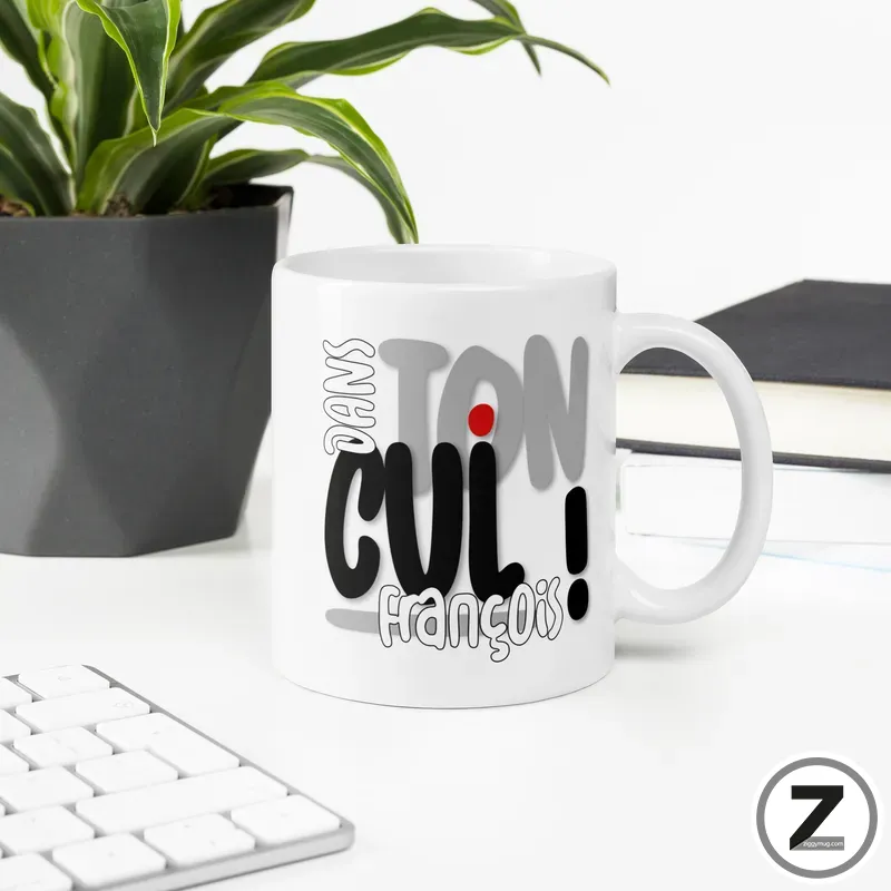 White mug with large black/gray graphic text on a white desk, next to a potted plant and notebook.