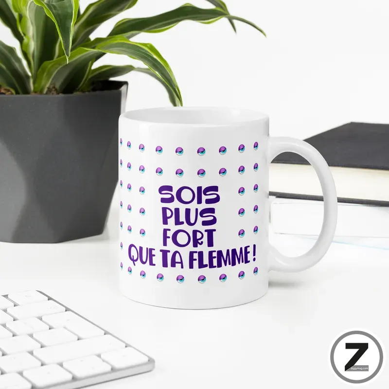 White ceramic mug on a desk featuring a purple phrase 'SOIS PLUS FORT QUE TA FEMME!' surrounded by small dotted motifs; a plant and keyboard are in the background.