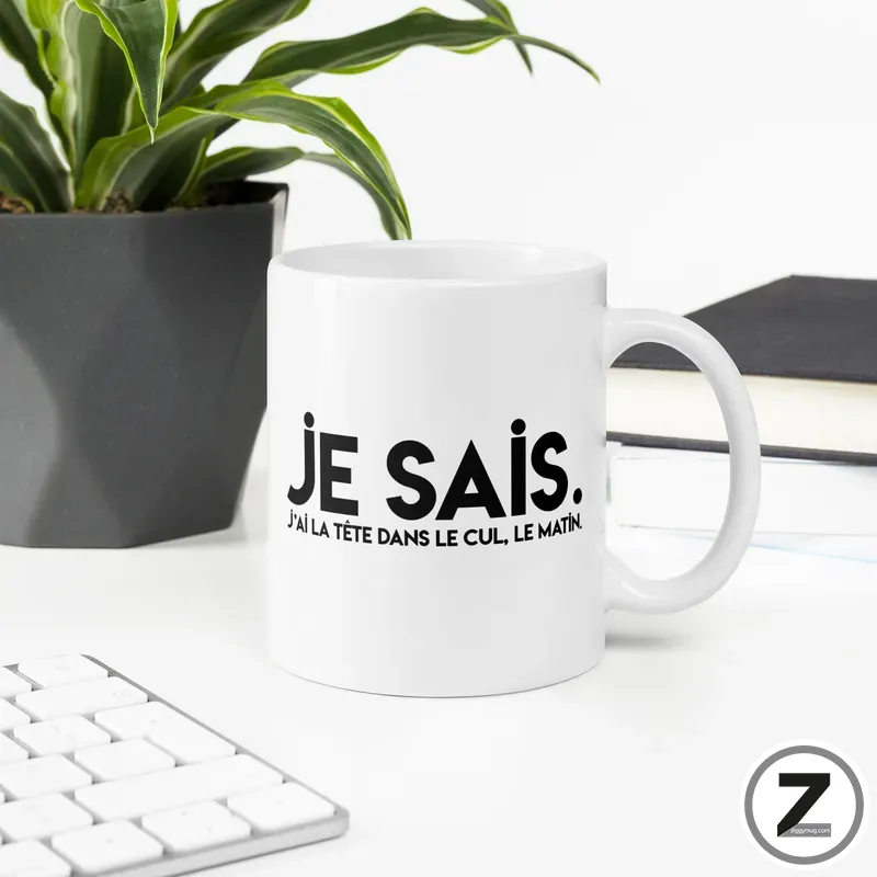 White coffee mug on a desk with a bold 'JE SAIS.' print and a small French phrase beneath; plant, keyboard, and books visible in the background.