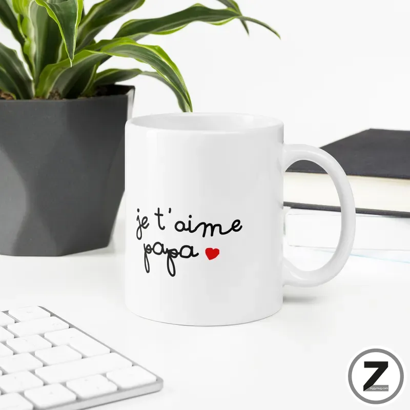 White ceramic mug on a white desk with handwritten 'je t'aime papa' and a red heart, beside a plant and keyboard.
