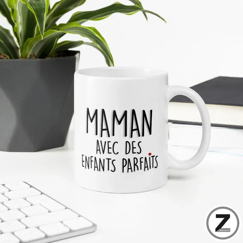White mug with the French phrase 'MAMAN AVEC DES ENFANTS PARFAITS' and a small red heart, on a desk with a plant and keyboard nearby.