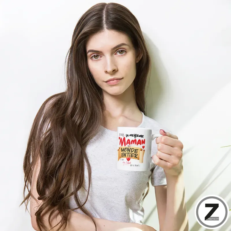 Young woman with long brown hair wearing a light gray T-shirt, holding a white mug with red and black text that reads 'Maman du monde entier' against a white background.