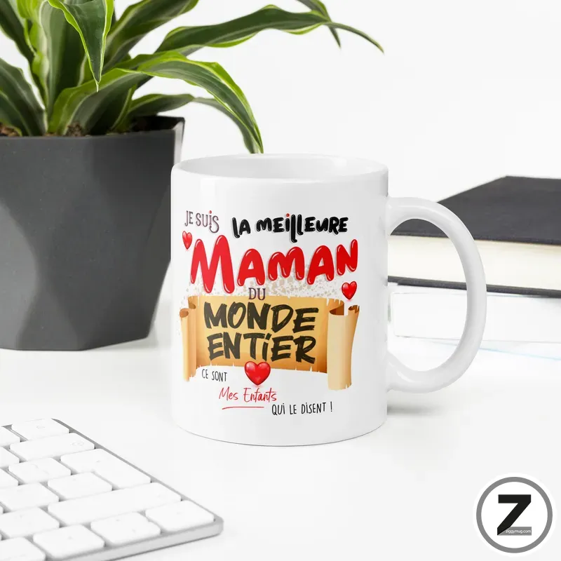 White mug on a desk featuring a red-and-black French message celebrating moms: 'Je suis la meilleure maman du monde entier' with hearts, behind a plant and keyboard.