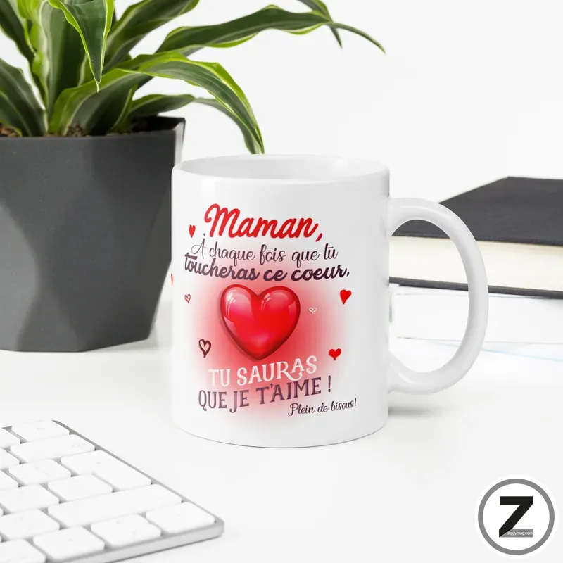 White ceramic mug with a red heart and a French loving message for Mom, placed on a desk beside a plant and a keyboard.
