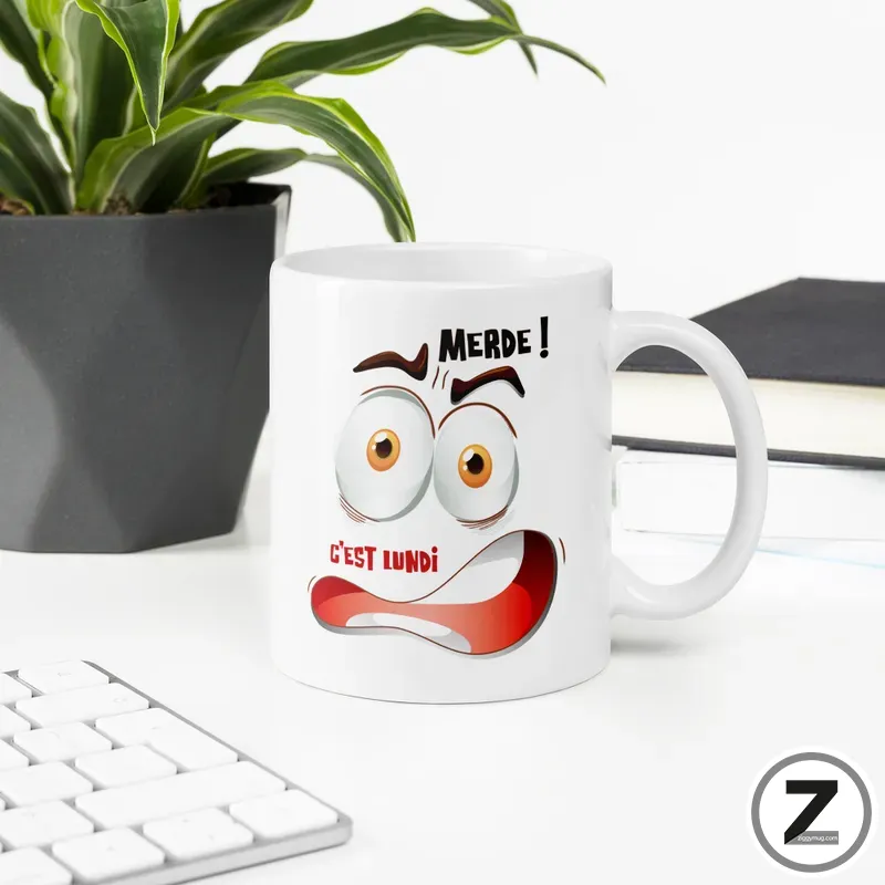 White mug with a cartoon surprised face and the French phrases 'MERDE!' and 'C'EST LUNDI' on a desk with a plant in the background and a keyboard nearby.