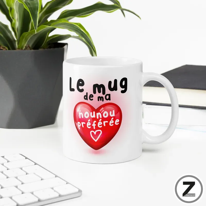 White ceramic mug with a large red heart design and French text on the heart, placed on a white desk beside a plant and keyboard.