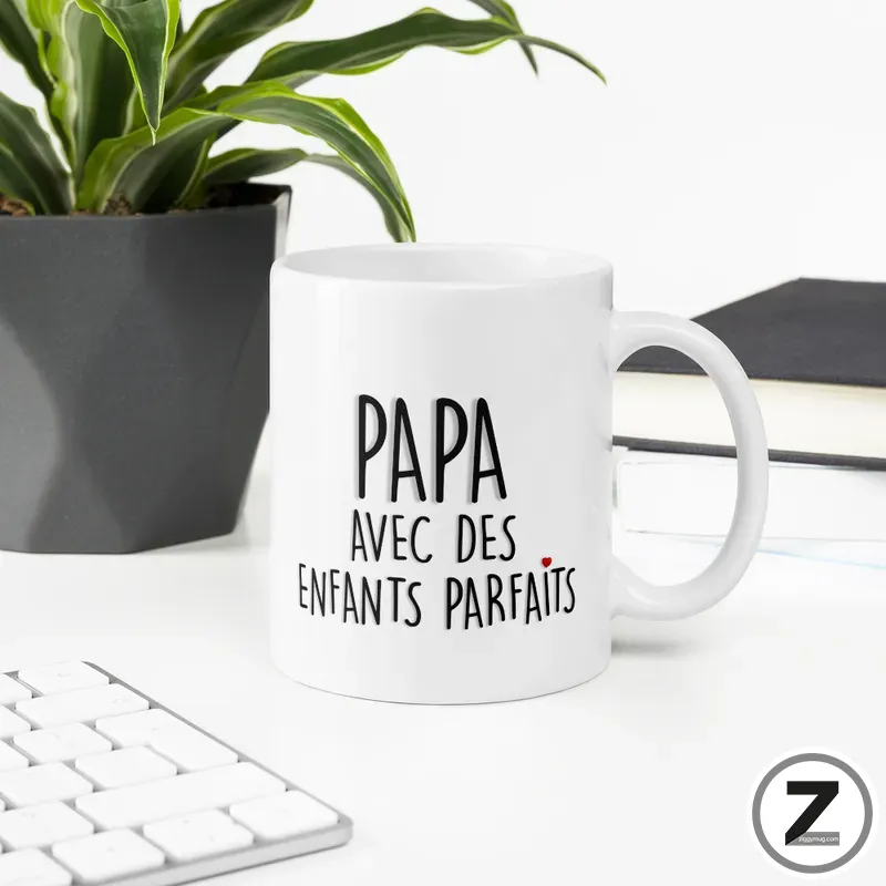 White mug on a desk beside a potted plant; mug reads 'Papa avec des enfants parfaits' in black letters on white ceramic.