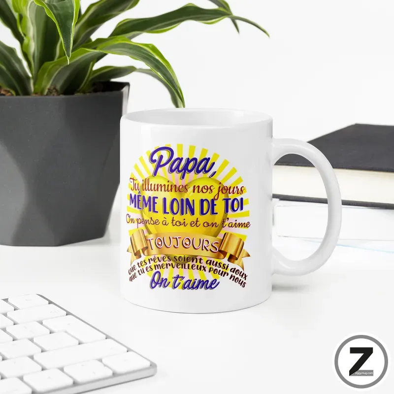 White ceramic mug on a desk featuring a colorful French message for dad, with a yellow sunburst design.