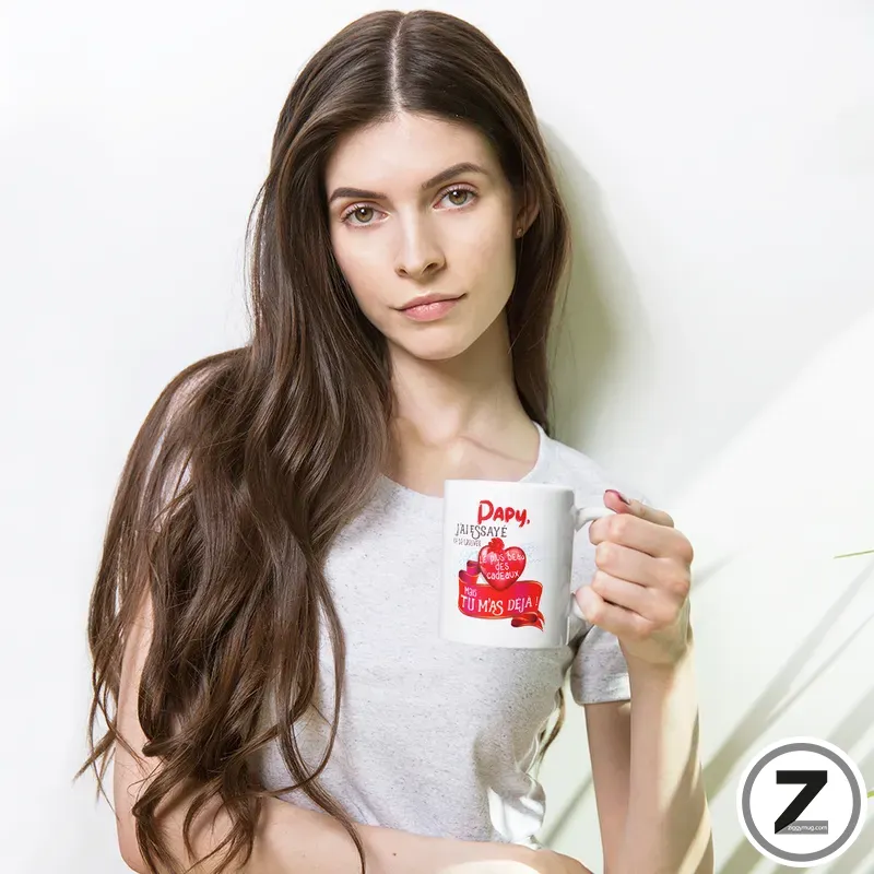 Woman with long brown hair wearing a light gray T-shirt, holding a white mug with red heart graphics and text, looking at the camera.
