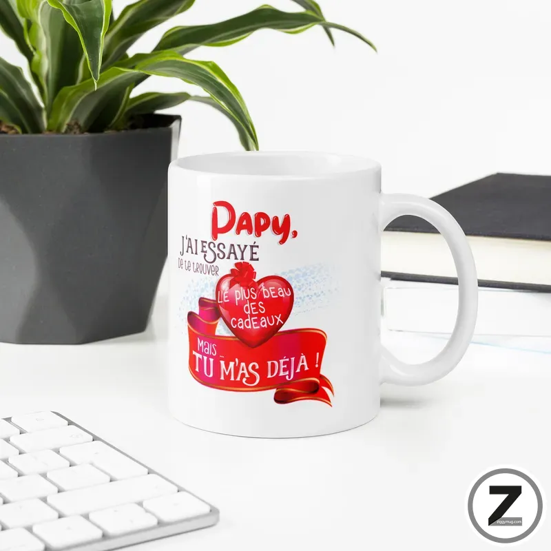 White mug with a red heart and ribbon graphic on a desk, featuring French text; a potted plant and books are in the background.