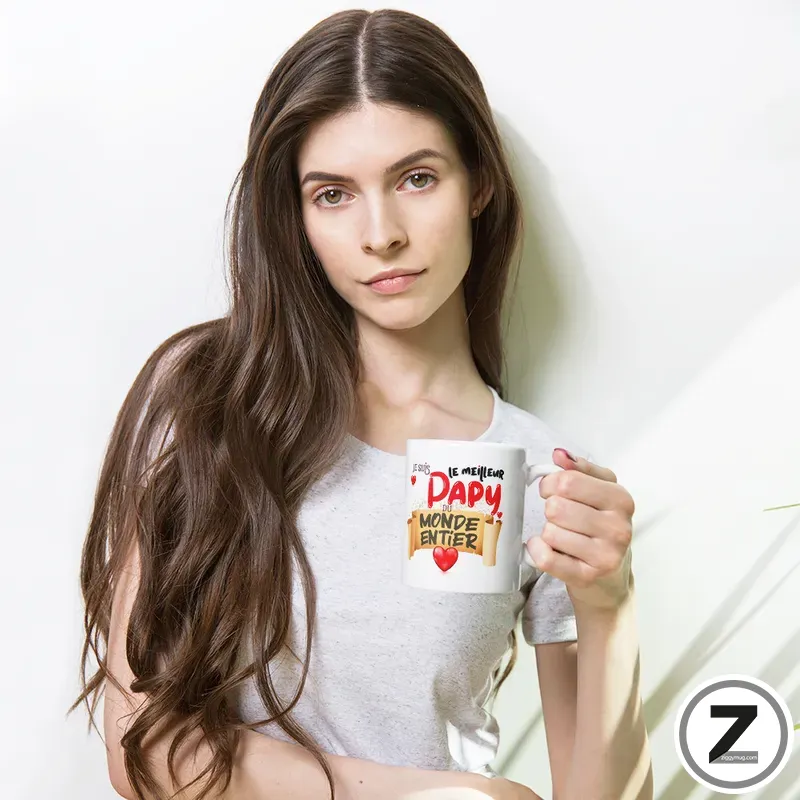 Young woman with long brown hair in a gray shirt holds a white mug with red lettering and hearts, standing against a light wall.