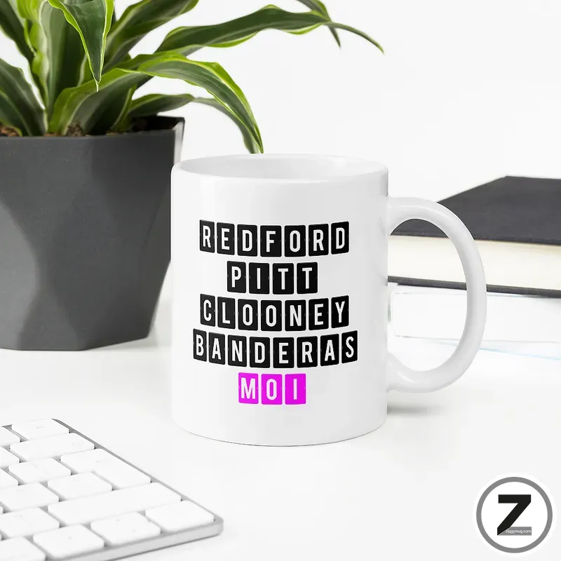 White ceramic mug on a desk features a black block-letter design with names Redford, Pitt, Clooney, Banderas, and 'moi' in pink at the bottom.