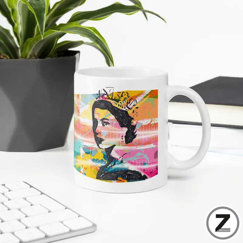 White ceramic mug with a colorful abstract portrait on its side, sitting on a white desk beside a plant.