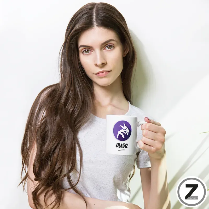 Young woman with long brown hair in a gray tee holding a white mug with a purple Judo logo, looking at the camera.
