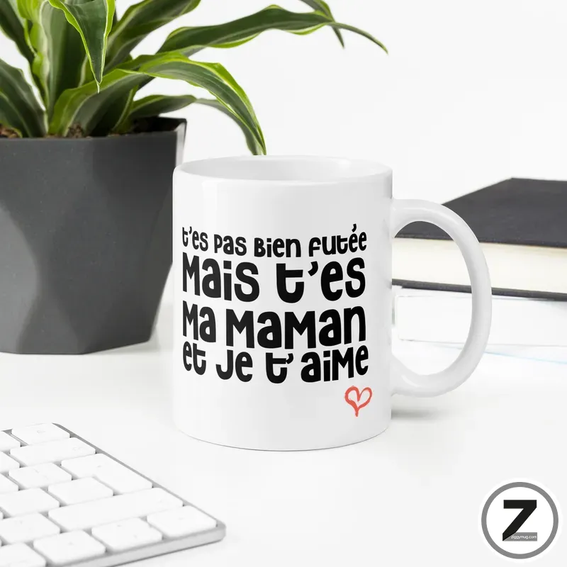 White mug on a white desk with bold French text about mom and love, plus a small red heart; plant and keyboard in the background.