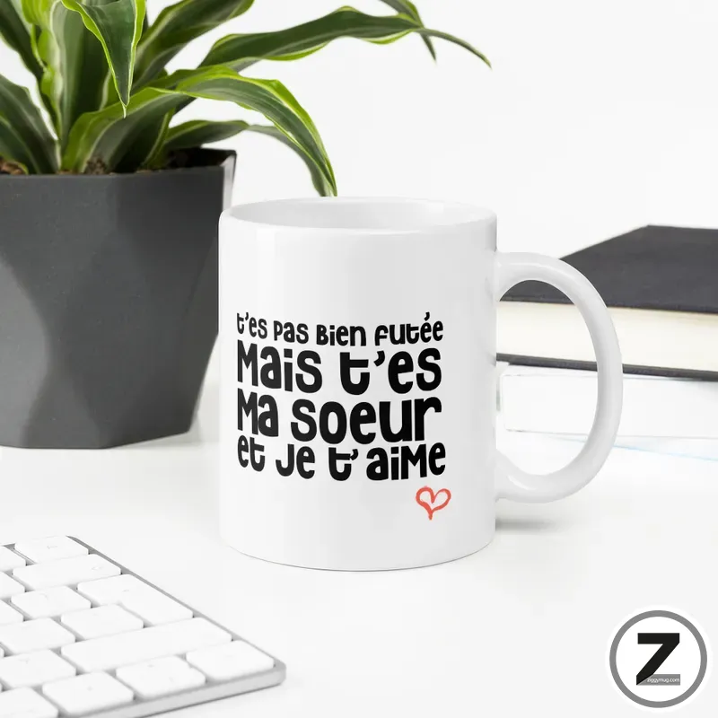 White mug with bold French text: "t’es pas bien foutée Mais t’es Ma sœur et je t’aime" and a red heart, on a desk with a plant and keyboard in the background.