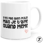 White mug with bold French phrase 't'es pas bien malin mais je t'aime quand même' and a small pink heart ornament.