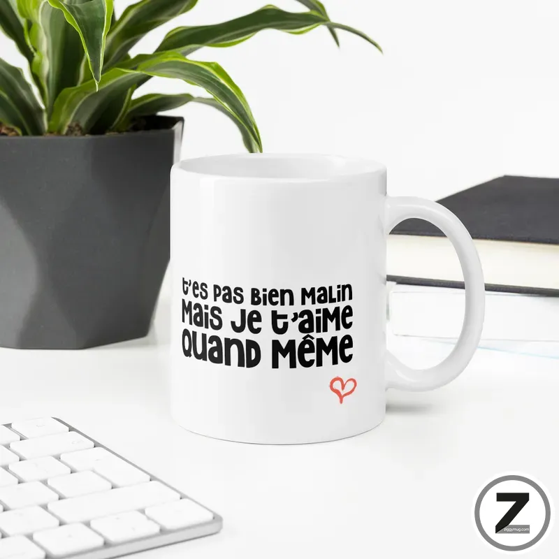 White ceramic mug with a black French phrase 't'es pas bien malin mais je t'aime quand même' and a small red heart, on a desk with a plant nearby.