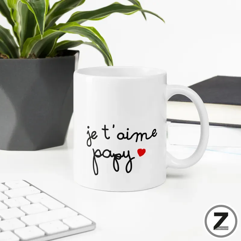 White ceramic mug on a desk with the text 'je t'aime papa' and a small red heart, beside a plant and keyboard.