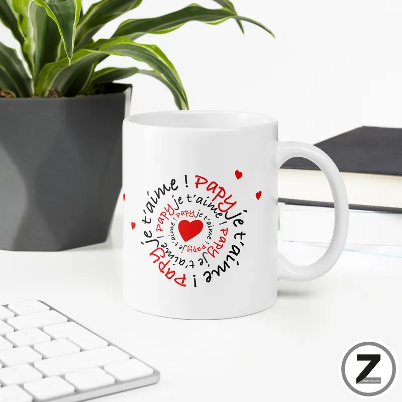 White ceramic mug with a red-and-black circular 'Je t'aime papa' design and small hearts, placed on a desk beside a plant.