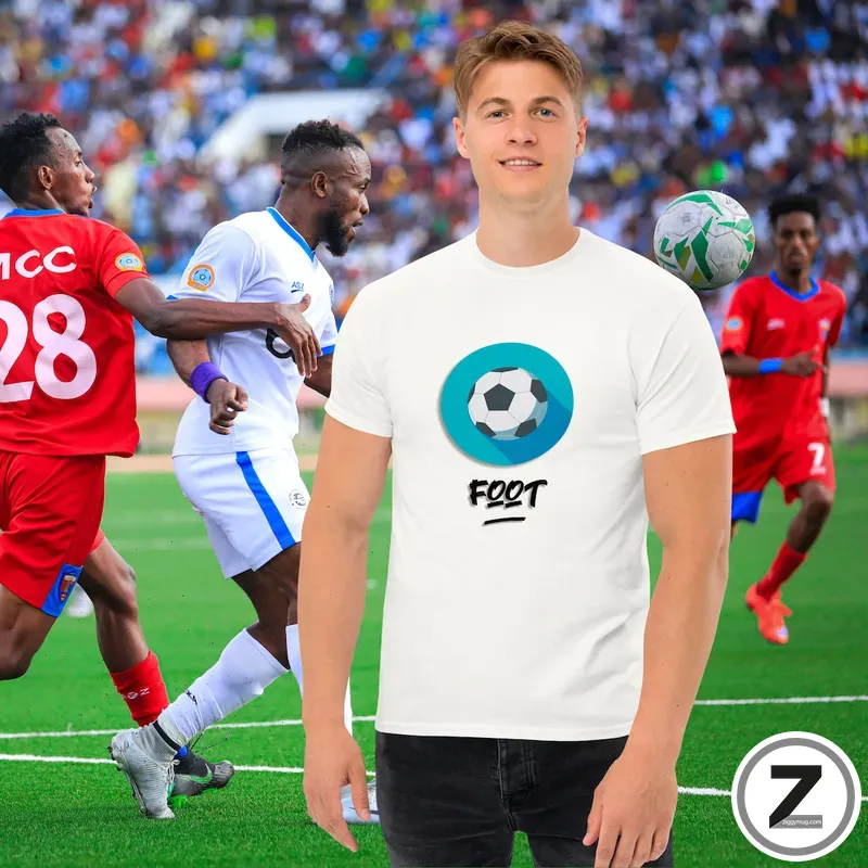 A man wearing a white t‑shirt with a blue-ringed soccer ball graphic stands on a sunny soccer field, with players in red and white jerseys racing in the background.